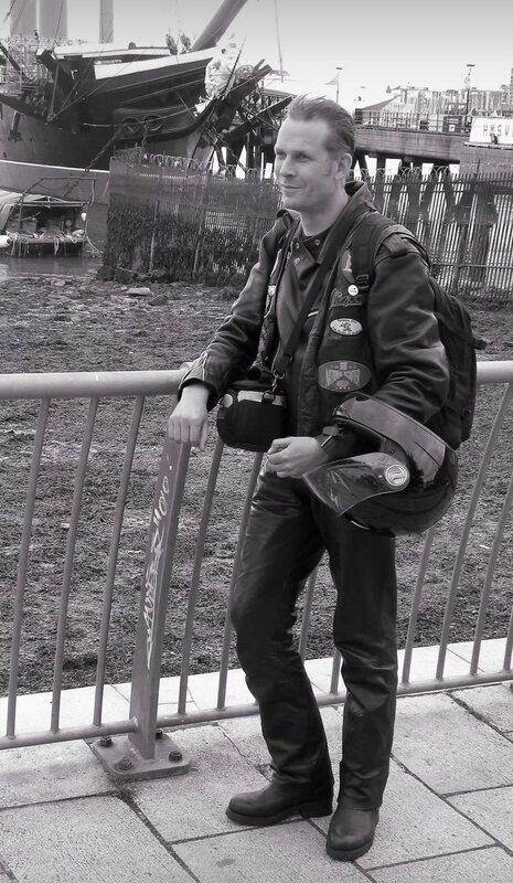 Stefan in Lederkombi mit Motorradhelm an einem Geländer am Hafen, im Hintergrund ein historisches Segelschiff, Schwarzweiß
