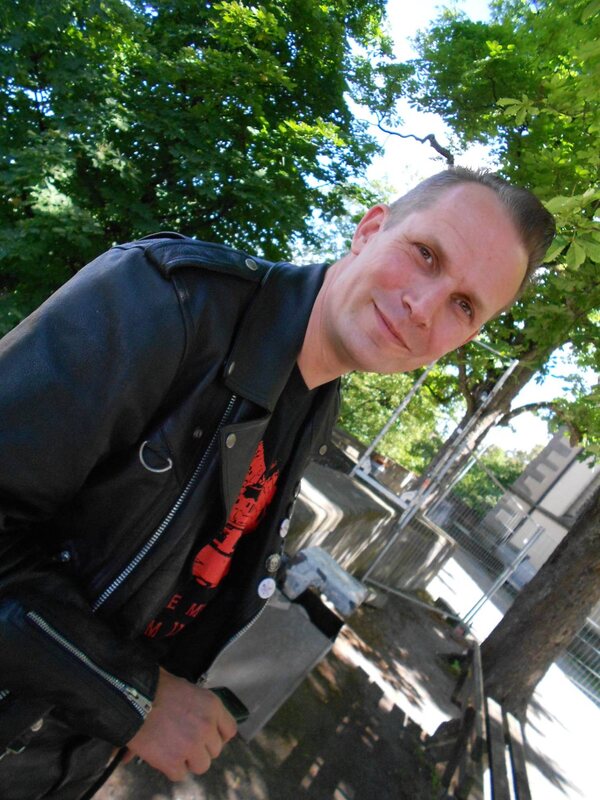 Stefan in Lederjacke, lächelnd und zur Kamera geneigt, unter grünen Bäumen